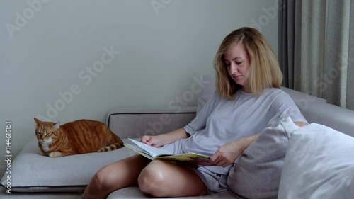 Young adult woman sits comfortably on couch absorbed in book while her cat lounges nearby. In living room caucasian female enjoys book with calm cat resting beside her reading and relaxation at home.