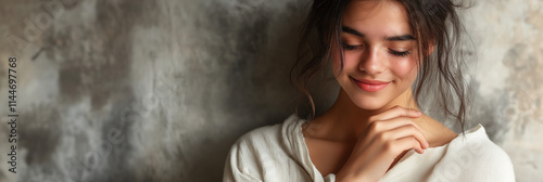 A young woman with a gentle smile looks downwards. Her dark hair frames her face, and she is wearing a soft sweater against a textured gray wall.