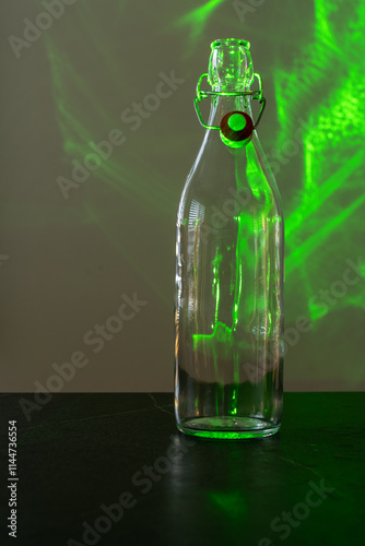 Clear swing top bottle is standing on a dark table, in front of a white background. A green laser is creating green textures on the bottle and the background. Vertical shot. photo