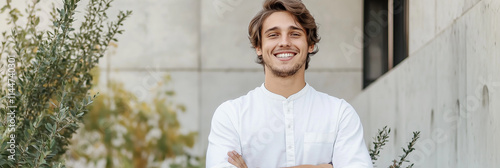 A young man with an affirming smile stands outdoors, exuding confidence and optimism amidst a minimal architectural background and fresh greenery. photo