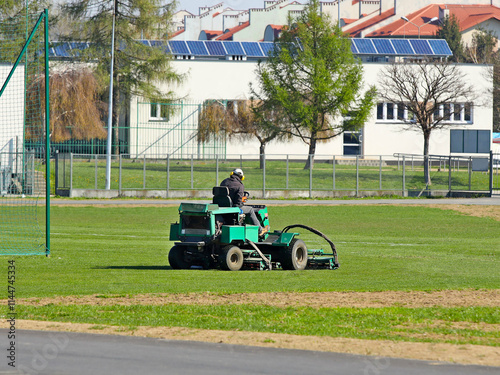 Worker on a large green lawn mower mows the grass on the football field. Landscape design and maintenance of green areas of the sports complex. Making grass stadium before the match. Human labor