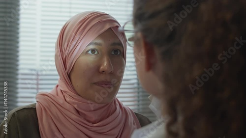 Close up shot of young female patient in headscarf being examined by young Caucasian female doctor in sterile gloves checking pupil reflex with diagnostic penlight in clinic photo