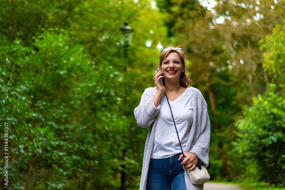 Beautiful middle aged woman walking and talking on phone against background of greenery in city park. Mobile phone conversation in park. Front view Beautiful middle aged woman walking and talking on phone against background of greenery in city park. Mobile phone conversation in park. Front view
