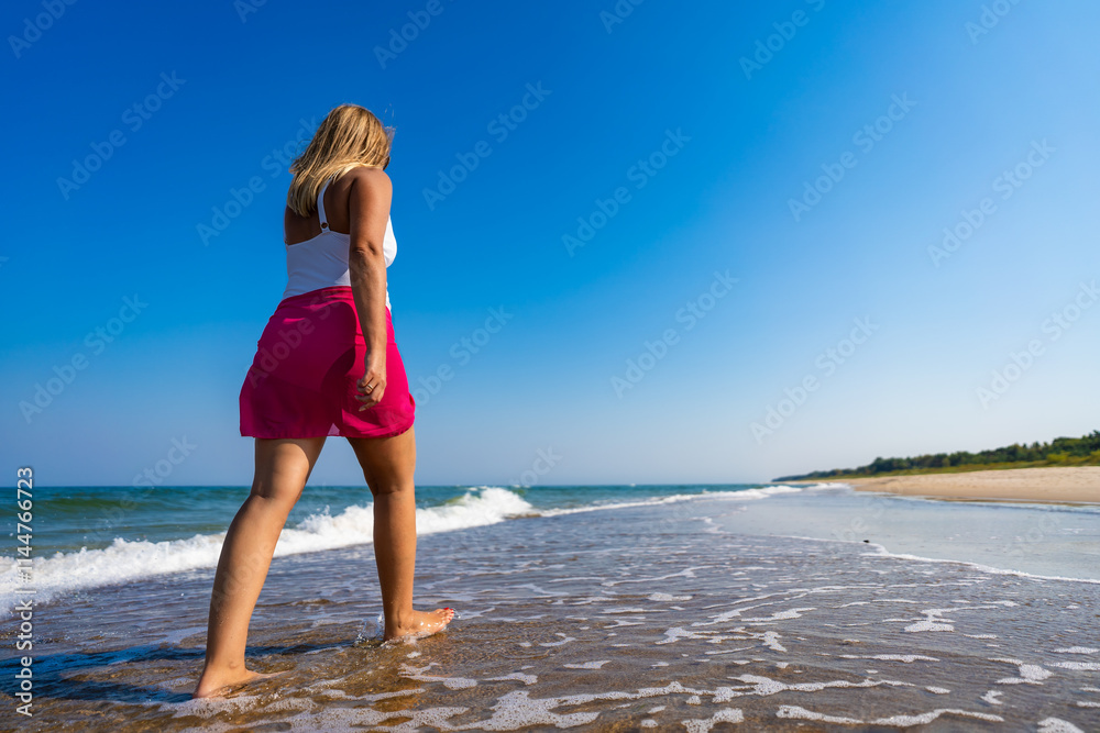 Naklejka premium Summer vacations. Beautiful mature woman walking on sandy beach on beautiful sunny day. Back view
