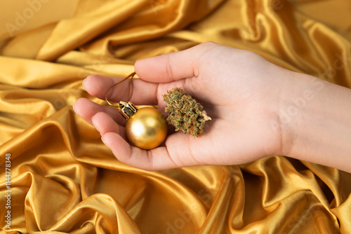 in white young female hand dry bud of medical marijuana and Christmas ball. On festive golden background