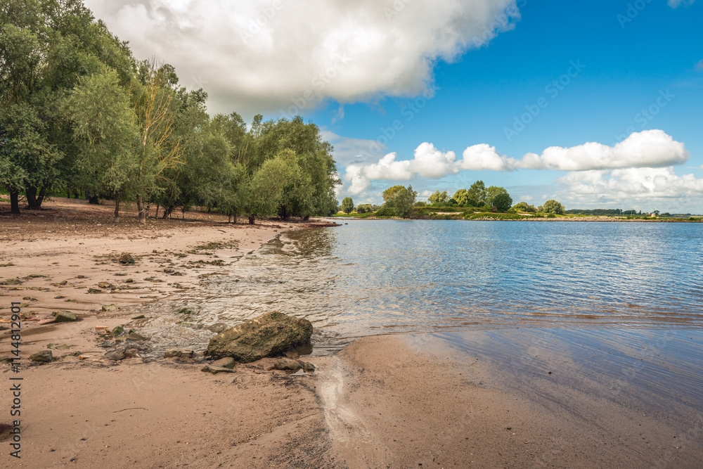 Picturesque landscape on the bank of a wide Dutch river. On the sandy beach there are a few stones. It is a cloudy day in the summer season with white clouds in the blue sky.