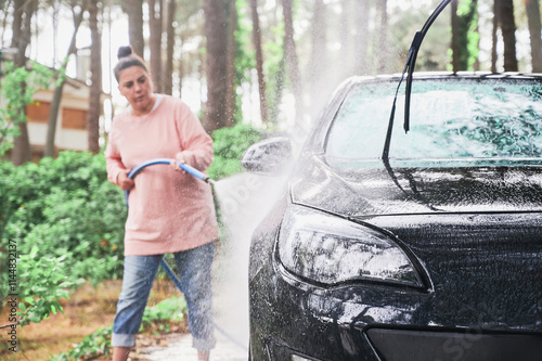 Black car is cleared by young woman using hose to spray water on driveway of her home