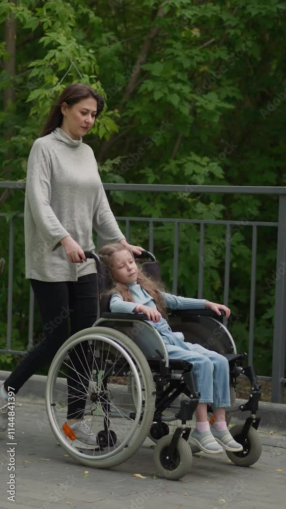 Mom walks on pavement with little daughter sleeping in wheelchair. Adult woman takes girl with muscular dystrophy out to city park on spring day Mom walks on pavement with little daughter sleeping in wheelchair. Adult woman takes girl with muscular dystrophy out to city park on spring day