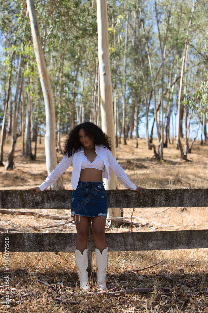 young, beautiful, Latin, brunette woman with curly hair dressed in white shirt, denim skirt, white boots and hat, leaning on the fence of the eucalyptus forest looking sensually at the camera. young, beautiful, Latin, brunette woman with curly hair dressed in white shirt, denim skirt, white boots and hat, leaning on the fence of the eucalyptus forest looking sensually at the camera.