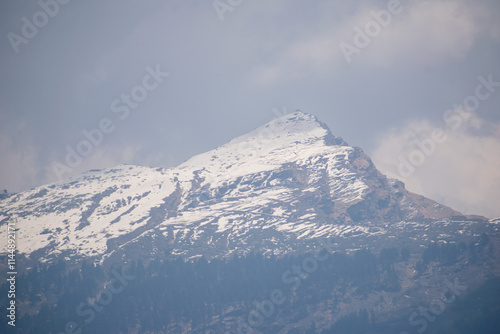 Chandrashila Peak, standing at 3690m in Uttarakhand, India, showcases a majestic snow-covered summit against a dramatic sky. The rugged slopes and forested base create a stunning Himalayan landscape. photo