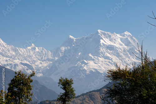 Majestic snow capped Himalayan peak Mt. Chaukhamba dominate the scene under a clear blue sky. Green trees and distant hills add depth to the landscape, creating a serene and breathtaking natural view. photo