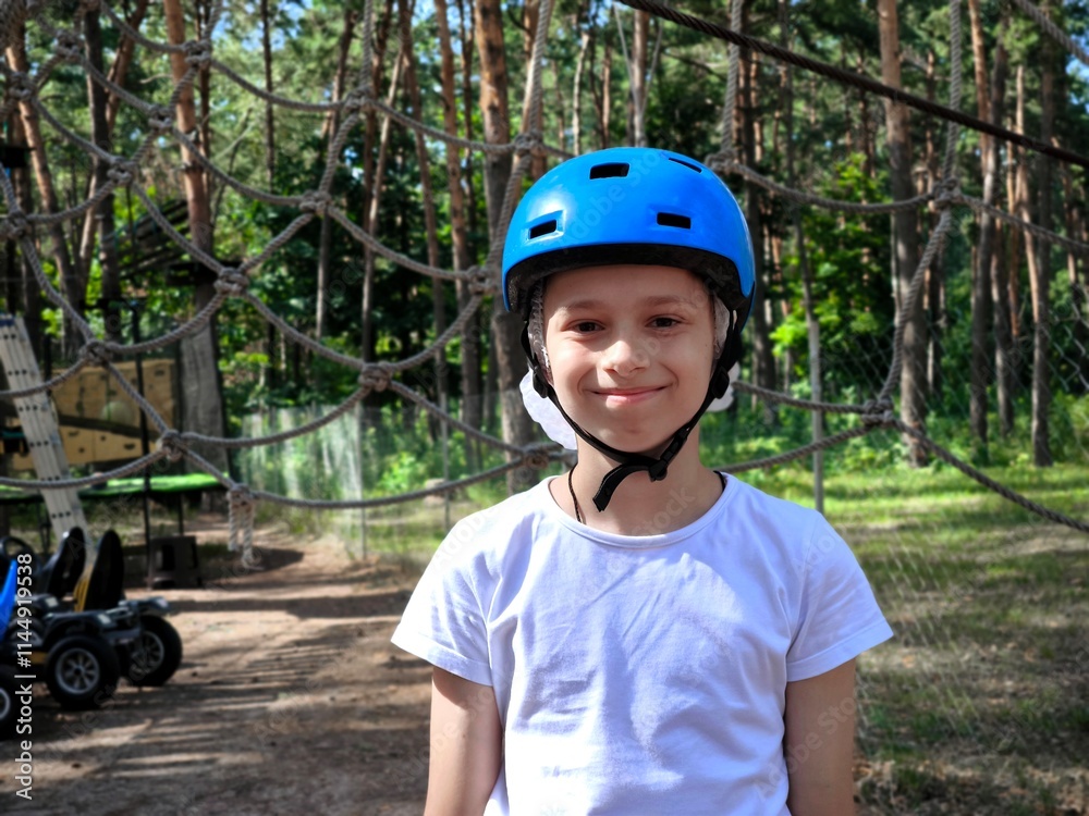 A child in a forest adventure park. A kid in a blue helmet and a white T-shirt is climbing a cable car. A children's entertainment center in an outdoor park. A child in a forest adventure park. A kid in a blue helmet and a white T-shirt is climbing a cable car. A children's entertainment center in an outdoor park.