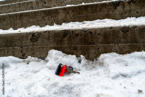 A set of keys left on snow-covered concrete stairs, suggesting an accidental drop or forgetfulness. The red key fob stands out vividly against the muted tones of the snow and worn concrete steps. 