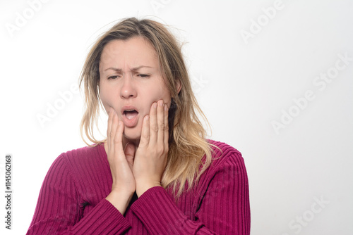 A woman holds her jaw with both hands, her face showing discomfort and worry. She appears to be experiencing pain or discomfort in her teeth or jaw area against a plain background.