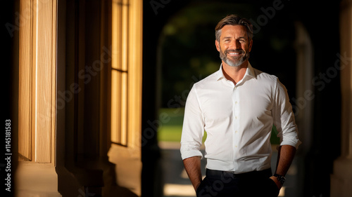 A confident man in a white shirt stands in a sunlit corridor, exuding charisma and professionalism, framed by classical architecture and warm light. photo