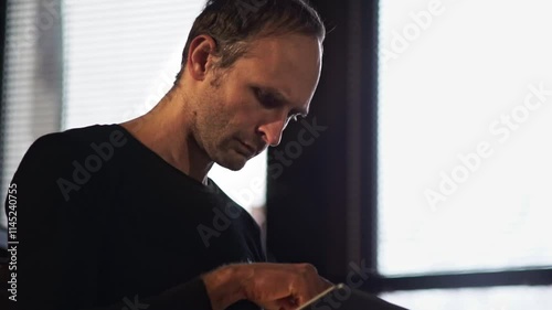 A man concentrates on his digital tablet, illuminated by natural light from a nearby window. The scene suggests a blend of technology use with a moment of quiet focus in a modern setting