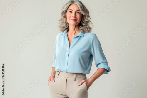 Half-length portrait of a woman in her 50s looking into the camera and wearing a casual blouse and trousers, standing against a solid-colored background