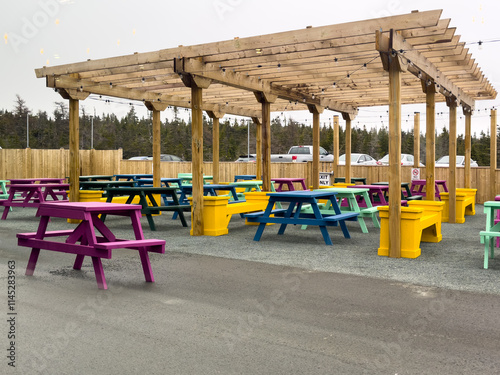 Wallpaper Mural A beer garden with multiple wooden picnic tables under a wooden shelter for sun protection. The benches and tables are painted colorful and bright paint. The backyard garden is enclosed with a fence. Torontodigital.ca