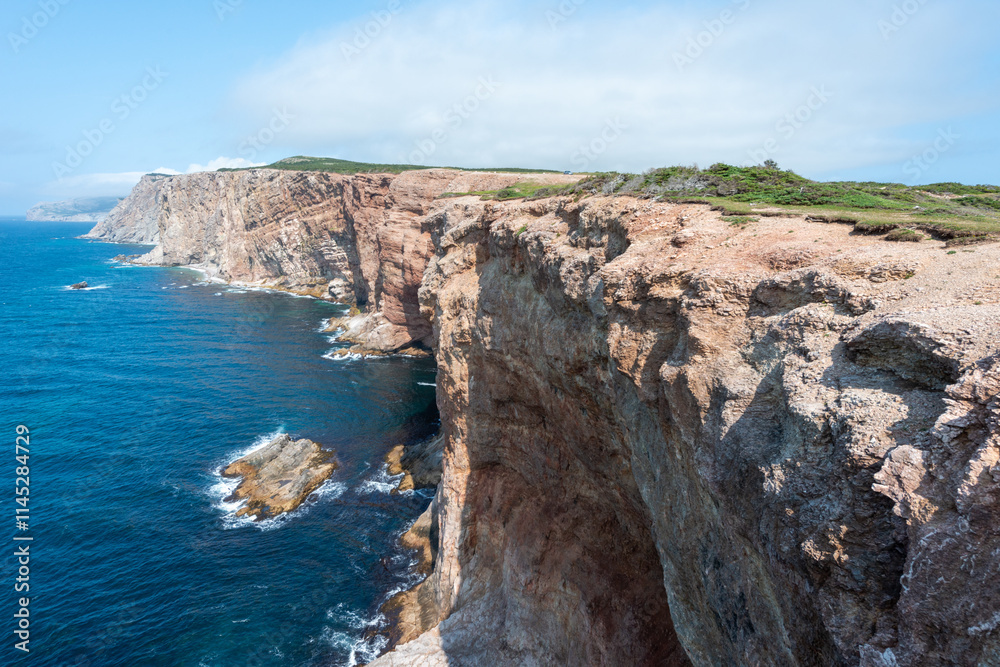 Near a deep blue ocean, a rugged coastline and cliff are in the foreground. It's a bright sunny day with a blue sky and a few clouds. An island with greenery, buildings, and rock is in the back. Near a deep blue ocean, a rugged coastline and cliff are in the foreground. It's a bright sunny day with a blue sky and a few clouds. An island with greenery, buildings, and rock is in the back.
