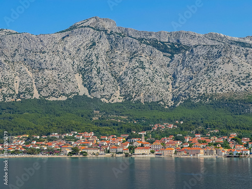 Orebic, Croatia - June 30, 2024: Church of Mary help of Christians in Orebic towers over the town and small chapel on Adriatic Sea shoreline, all set in red roofed cityscape. Mountain range in back photo