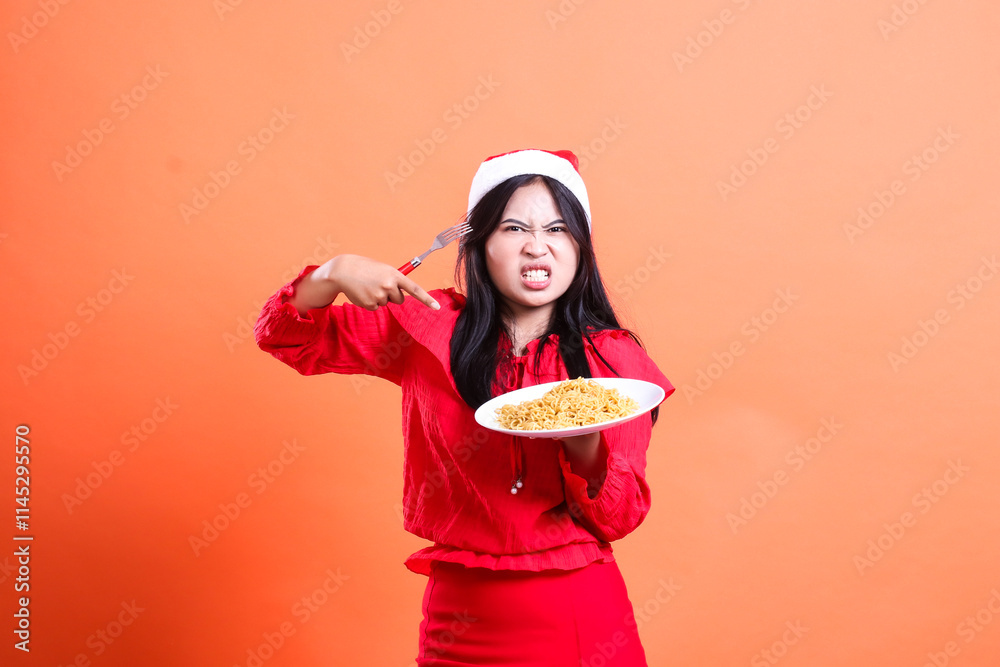 Asian female beauty annoyed looking at camera, hand holding fork and white plate filled with noodles while pointing at it, wearing red christmas blouse, isolated on orange background Asian female beauty annoyed looking at camera, hand holding fork and white plate filled with noodles while pointing at it, wearing red christmas blouse, isolated on orange background