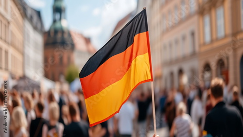 A German flag is waving in a crowd of people. City buildings are in the background.