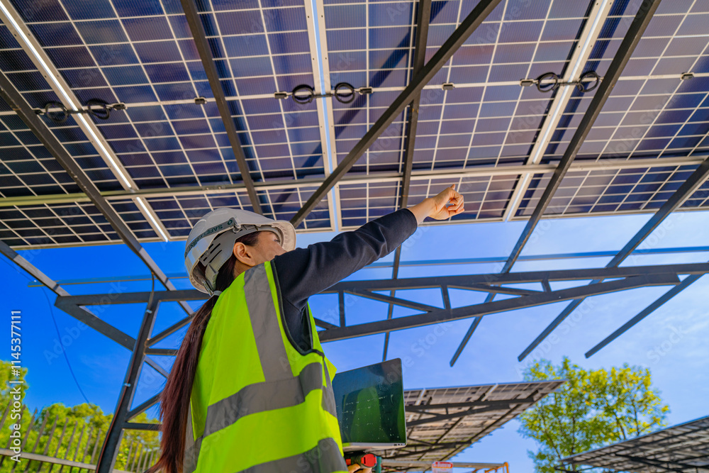 Fototapeta premium A technician in a safety helmet and reflective vest inspects a solar panel structure using a laptop. The solar panels are mounted on a steel carport framework under a clear blue sky.