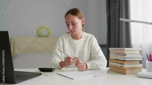 Concentrated teenage student girl sitting at desk writing notes in notebook, studying at home using laptop, books and calculator, doing homework preparing for exam