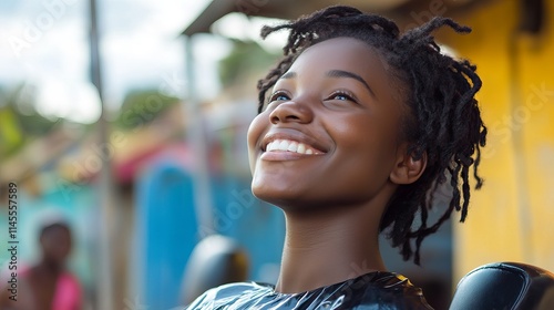 A young Black African woman sitting in an outdoor salon, enjoying her hair makeover, looks up at the sky with a bright smile. 