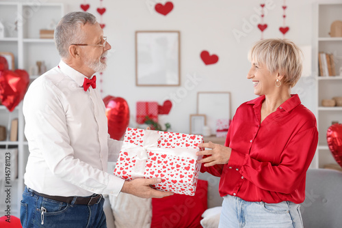 Mature man greeting his happy wife with gift box at home. Valentine's Day celebration