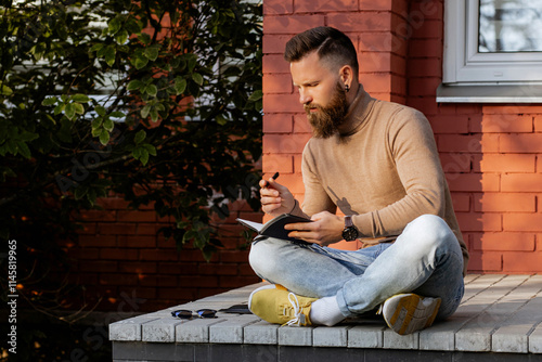 Stylish bearded man in brown turtleneck sweater and jeans sitting in lotus pose on beton steps outside holding sketchpad, painting tattoo sketches, writing notes. Male working remote photo