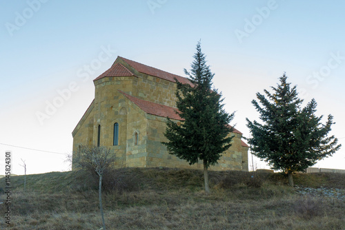 A small stone church with a tiled roof on a hill above the city of Tbilisi. Two fir trees and a tree without leaves. photo