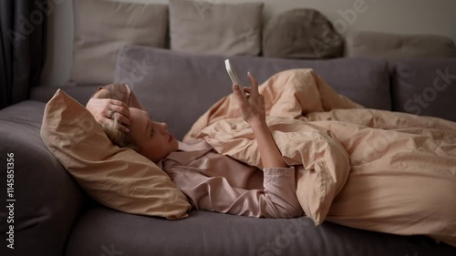 Zoom Out blonde girl in a pink nightie lies under an orange blanket and responds to social networks after waking up in the bedroom in the morning