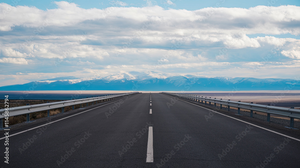 Fototapeta premium Vast Asphalt Road Stretching into the Horizon with Clear White Dividing Lines and Metal Guardrails, Surrounded by Majestic Snow-Capped Mountains, Fluffy Clouds in the Sky, Evoking Adventure and Explor