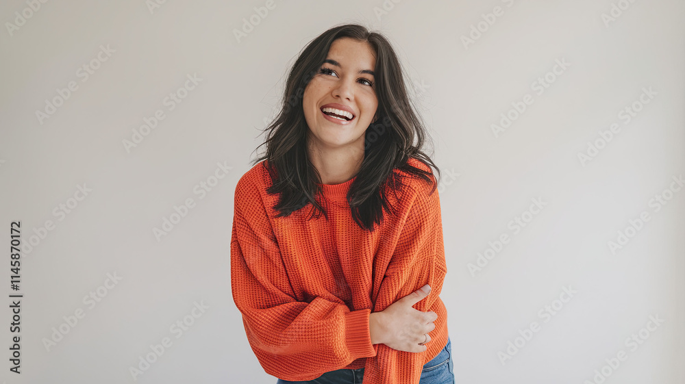 Young Woman with Dark Hair Wearing Bright Orange Sweater and Blue Jeans, Smiling Widely Against a White Background, Expressing Joy, Happiness, and a Moment of Accomplishment or Celebration Young Woman with Dark Hair Wearing Bright Orange Sweater and Blue Jeans, Smiling Widely Against a White Background, Expressing Joy, Happiness, and a Moment of Accomplishment or Celebration