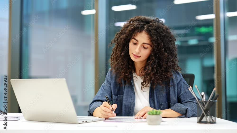 Focused businesswoman with curly hair works on laptop and makes notes at desk in modern office. Professional analyzes data, develops project strategy, and performs tasks. Focused businesswoman with curly hair works on laptop and makes notes at desk in modern office. Professional analyzes data, develops project strategy, and performs tasks.