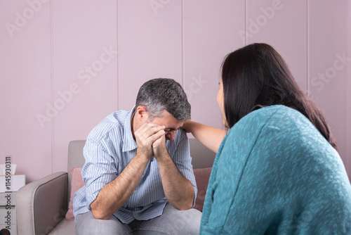 A man in psychological consultation cries with his hands covering his face, while the psychotherapist, with her back turned, accompanies him in a warm and professional environment. 