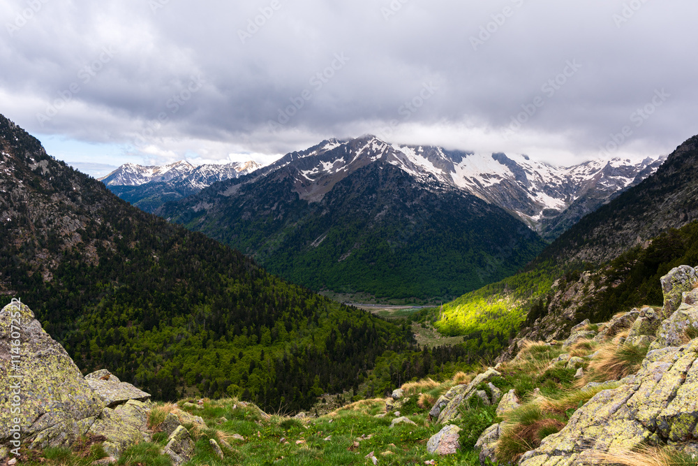 Fototapeta premium Paisaje alpino con montañas nevadas, cielo nublado y laderas verdes en primavera. Un rayo de sol ilumina una parte del bosque. Escenario natural impresionante y sereno. GR11, Lleida, Catalunya