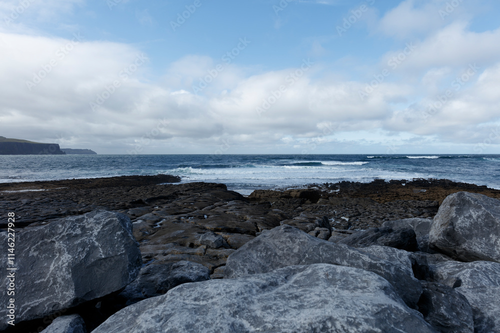 Rocky shoreline meets a vast expanse of ocean under a partly cloudy sky. Waves gently lap against the dark basalt rocks. Rocky shoreline meets a vast expanse of ocean under a partly cloudy sky. Waves gently lap against the dark basalt rocks.