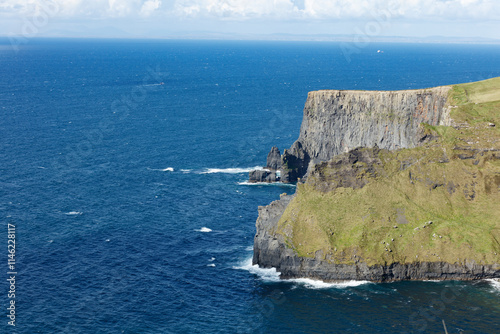 Dramatic coastal cliffs of the Cliffs of Moher, Ireland, meet the churning blue Atlantic Ocean.  Waves crash against the basalt rock face. photo