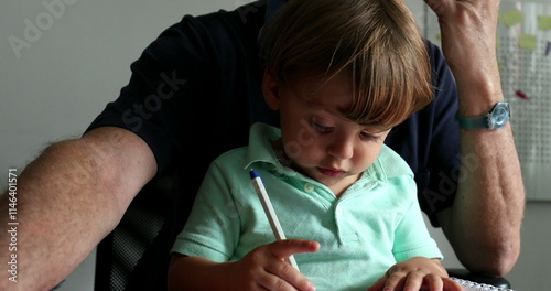 Multi-tasking grand-father working while taking care of toddler boy while he doodles photo