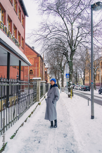 A woman stands on a snowy street, wearing a grey coat and colorful hat, capturing a serene winter moment.