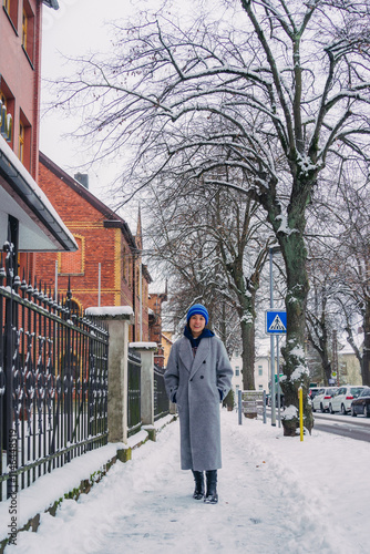 A woman stands on a snowy street, wearing a grey coat and colorful hat, capturing a serene winter moment.