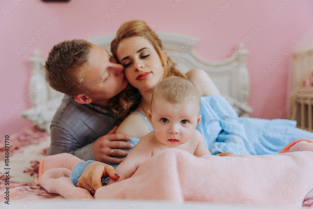 A man and a woman are kissing a baby on a bed. The baby is wrapped in a pink blanket A man and a woman are kissing a baby on a bed. The baby is wrapped in a pink blanket