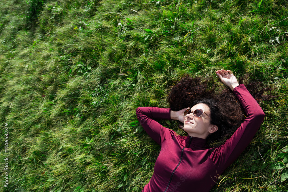 Curly haired woman wearing sunglasses lying down on a grass. Attractive cheerful lady relaxing outdoors. Copyspace Curly haired woman wearing sunglasses lying down on a grass. Attractive cheerful lady relaxing outdoors. Copyspace