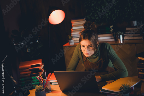 Young woman in a cozy brown jumper intently studying at her desk in a softly lit interior setting, surrounded by books and a lapto photo