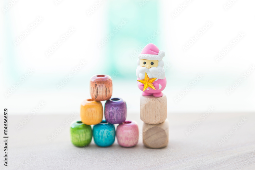 Fototapeta premium Christmas card background idea, colourful wooden beads stack with pink Santa Clause and star on wooden cube over blurred background, outdoor day light