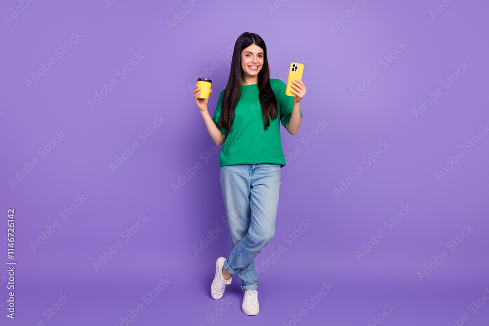 Fototapeta premium Young woman in green t-shirt with coffee cup and smartphone standing against a purple background, displaying a joyful and trendy expression