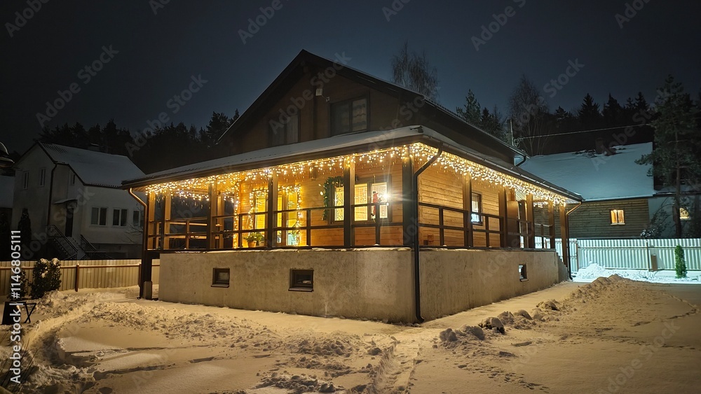 Obraz premium Garlands are lit on the walls and terrace of a wooden cottage in the evening darkness on Christmas Eve. Everything around is covered in snow. The path is cleared in snowdrifts. Nearby is a forest