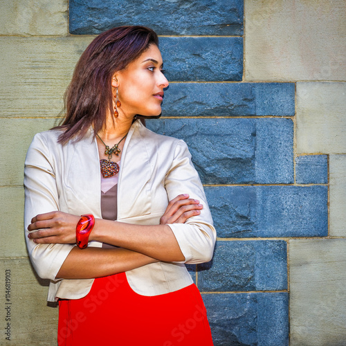A woman stands with her arms crossed, gazing to the side while leaning against a brick wall. She wears a beige jacket over a red dress and accessorizes with earrings, a necklace, and a bracelet.